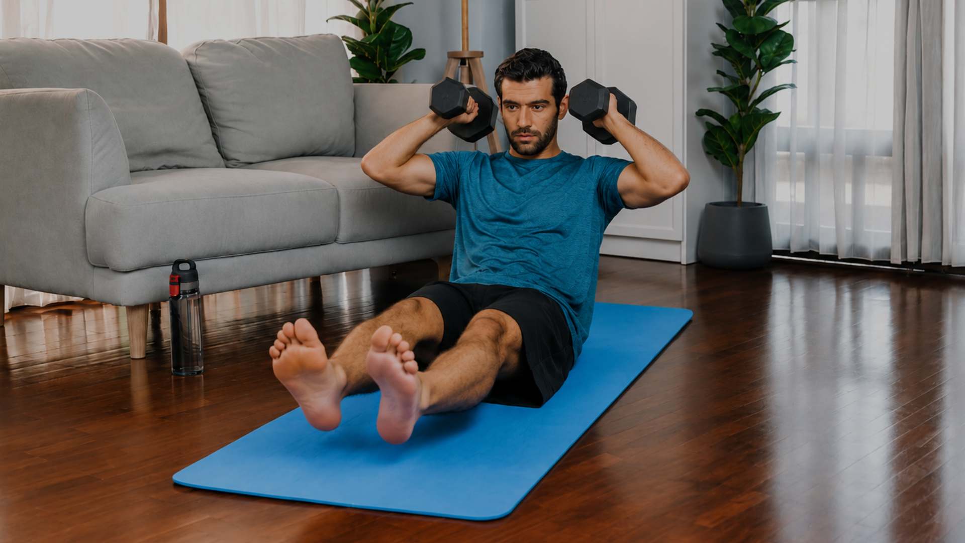 A man in a blue t-shirt and black shorts sits on a blue exercise mat in a living room, holding black dumbbells overhead. A gray sofa, wooden floor, and potted plants are visible in the background.