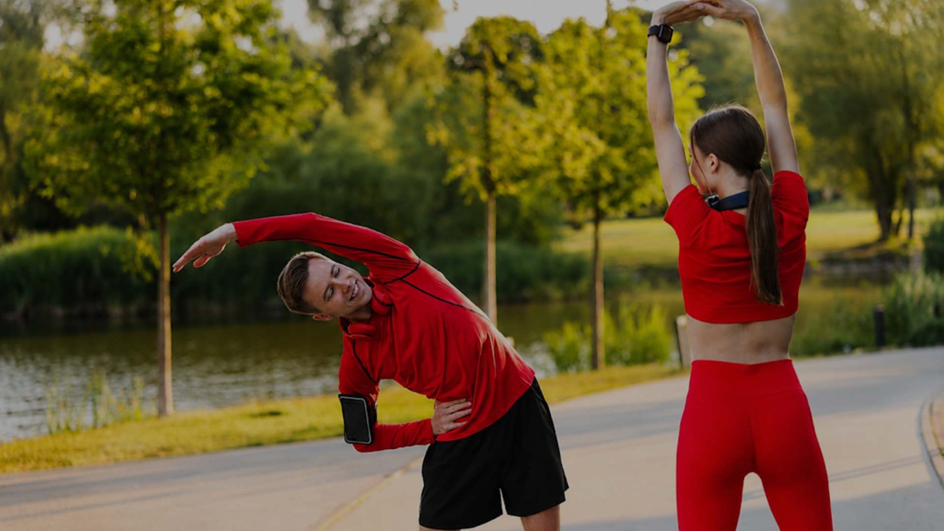A man and a woman in matching red activewear outfits stretch on a paved path next to a lake, with trees in the background.