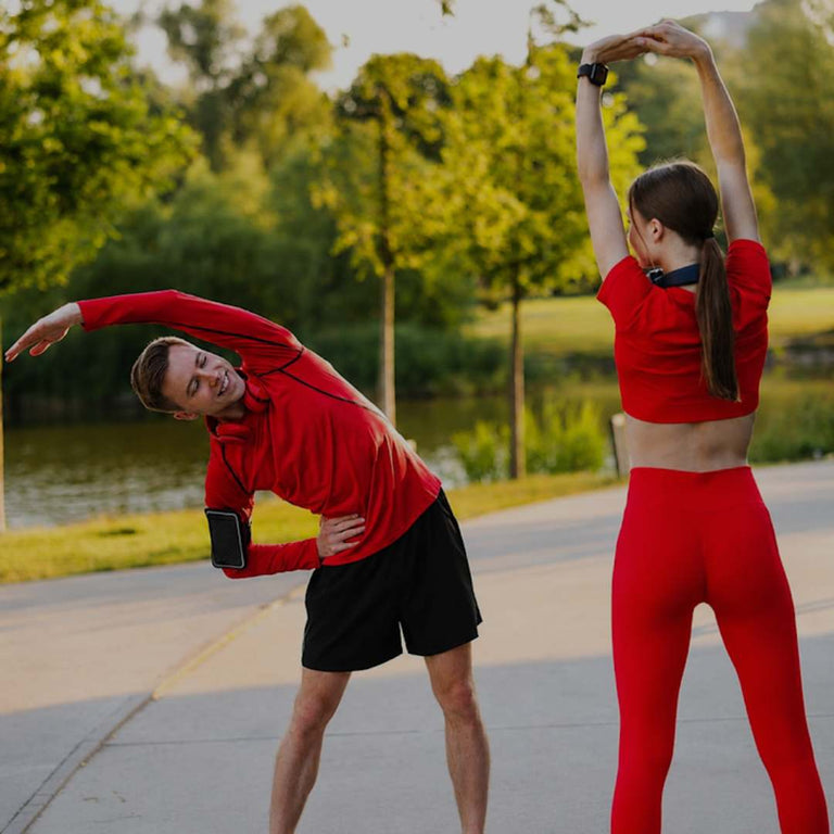 A man and a woman in matching red activewear outfits stretch on a paved path next to a lake, with trees in the background.